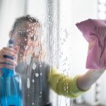 Young woman cleaning window at home using spray detergent and rag